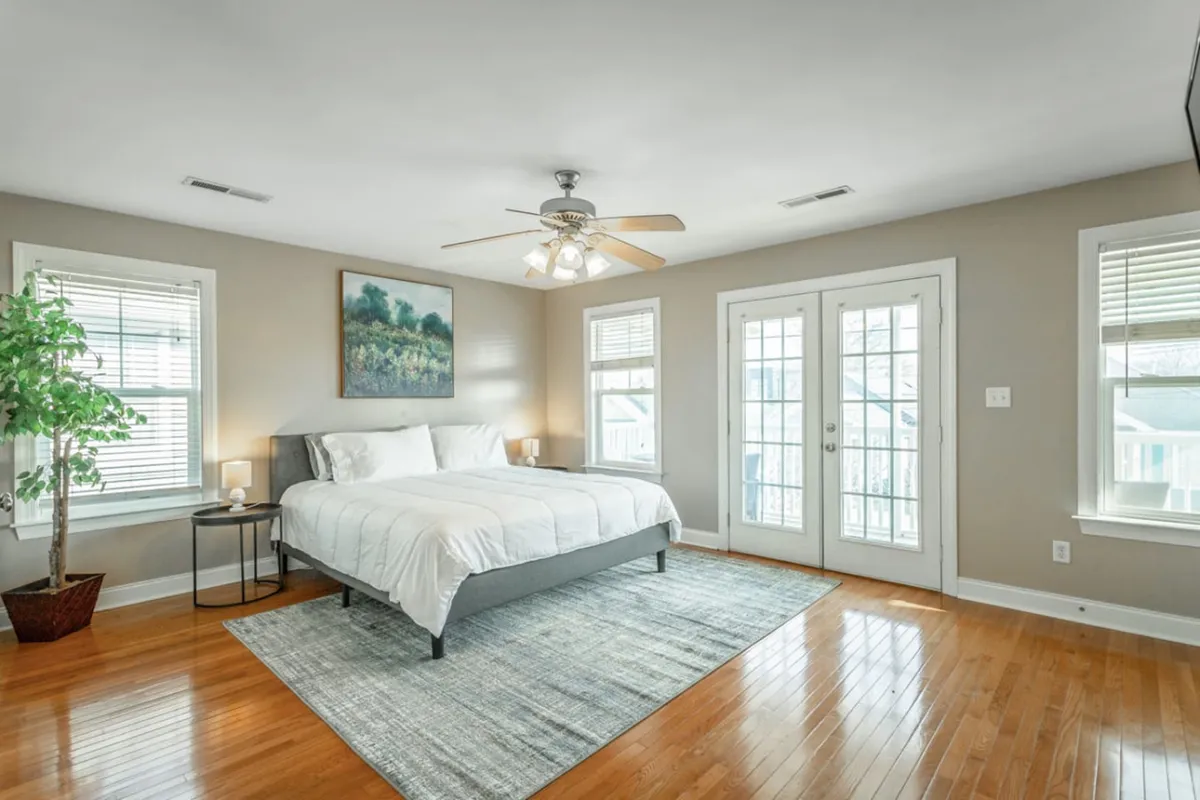 A tranquil bedroom featuring a neatly made bed with soft linens, stylish wall art, double doors that let in natural light, and a touch of greenery with a potted plant. The room feels spacious, airy, and perfectly suited for relaxation or a quiet read before bed.