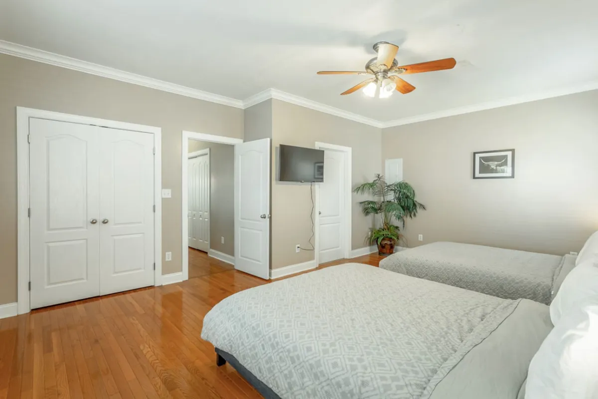 A cozy bedroom with two beds, each neatly arranged and inviting. The wall-mounted TV, indoor plant, and closet doors create a functional yet homelike atmosphere, ideal for both families and business travelers. The overall look is comfortable and uncluttered, with space for relaxation and storage.