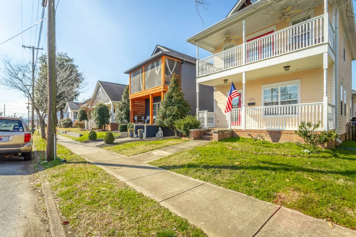 The image shows two neighboring houses with front porches adorned with an American flag and manicured lawns, exuding a sense of community and walkability. The friendly streetscape hints at a lively, accessible neighborhood environment.