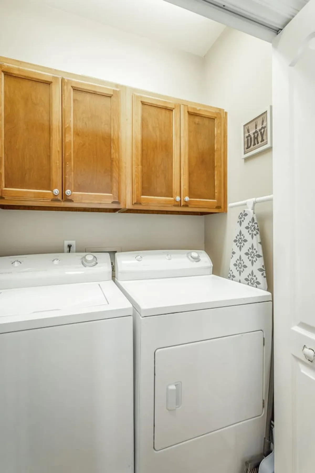 A bright, functional laundry area featuring a washing machine and dryer set beneath wooden cabinets, complemented by a decorative wall sign. The space is organized and practical, emphasizing ease of daily routines.