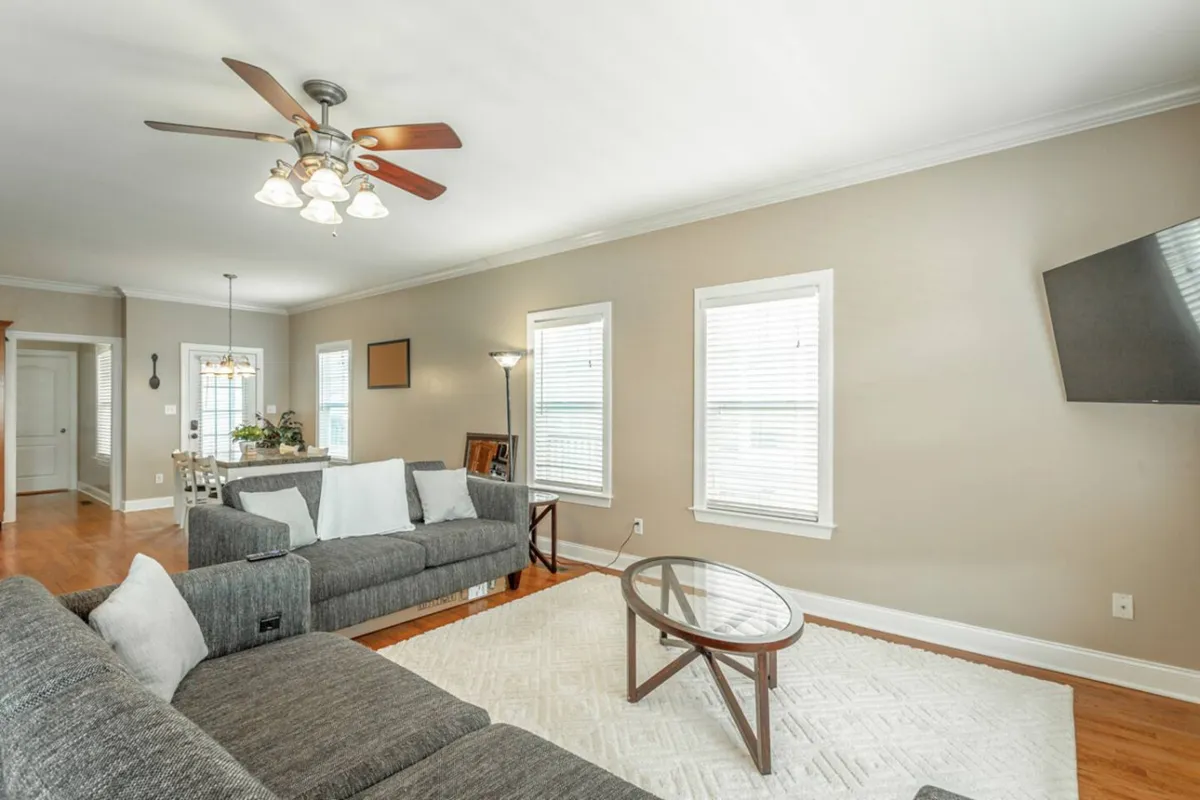 Showing a comfortable living area with a large sofa, coffee table, windows that let in abundant daylight, and a visible light fixture. The scene suggests a space perfect for socializing, relaxing with family, or enjoying a game night, all in a sunlit, airy environment.