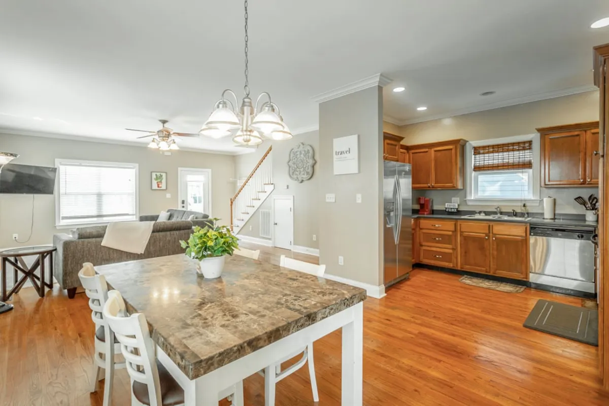 This image captures an open-plan layout featuring a dining table, kitchen area, staircase, and a glimpse of a cozy living area. The space is filled with natural light, and the arrangement invites both group gatherings and intimate meals. Plush seating and tasteful decor add to the warmth and comfort, aligning with the theme of togetherness and modern downtown living.