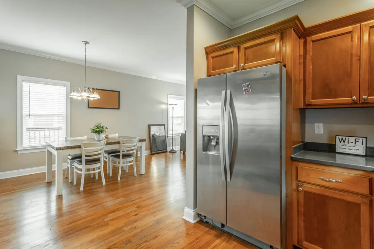 The image shows a cozy dining area adjacent to the kitchen, featuring a wooden dining table with matching chairs illuminated by natural light from a nearby window. Stainless steel appliances are visible, linking the dining and kitchen spaces for a seamless family or group dining experience.