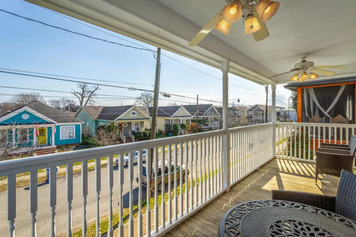 A porch railing with wicker furniture overlooks the street, offering a view that connects the home to the liveliness outside. The setting is cozy and inviting, perfect for relaxing and observing the neighborhood activity.